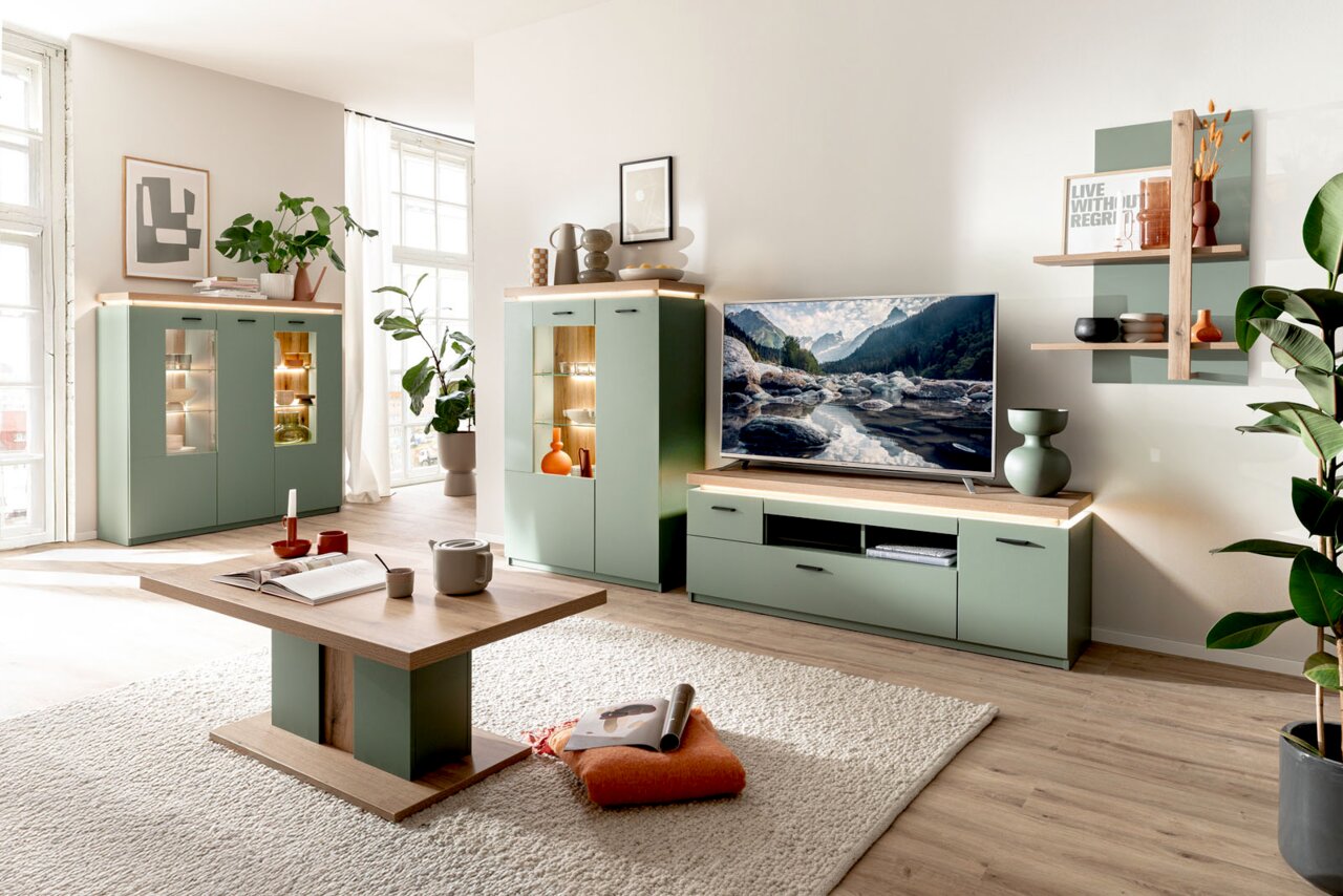 Modernes Wohnzimmer-Setup mit salbeigrüner Wohnwand (Vitrinen und TV-Lowboard mit Holzoberfläche und Beleuchtung), Couchtisch und Wandregal, fotografiert in frontaler Raumansicht mit leichter Perspektive von links.