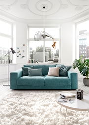 Zweiersofa in Türkis in einem hellen Wohnzimmer mit großen Fenstern, von vorne gesehen.