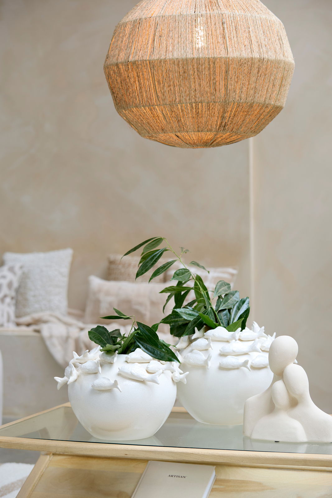 Zwei weiße Vasen mit Fischornamenten, gefüllt mit grünen Pflanzen, auf einem Glastisch. Im Hintergrund eine gemütliche Wohnzimmerszene mit Kissen und einer hängenden Lampe. Perspektive: seitlich.