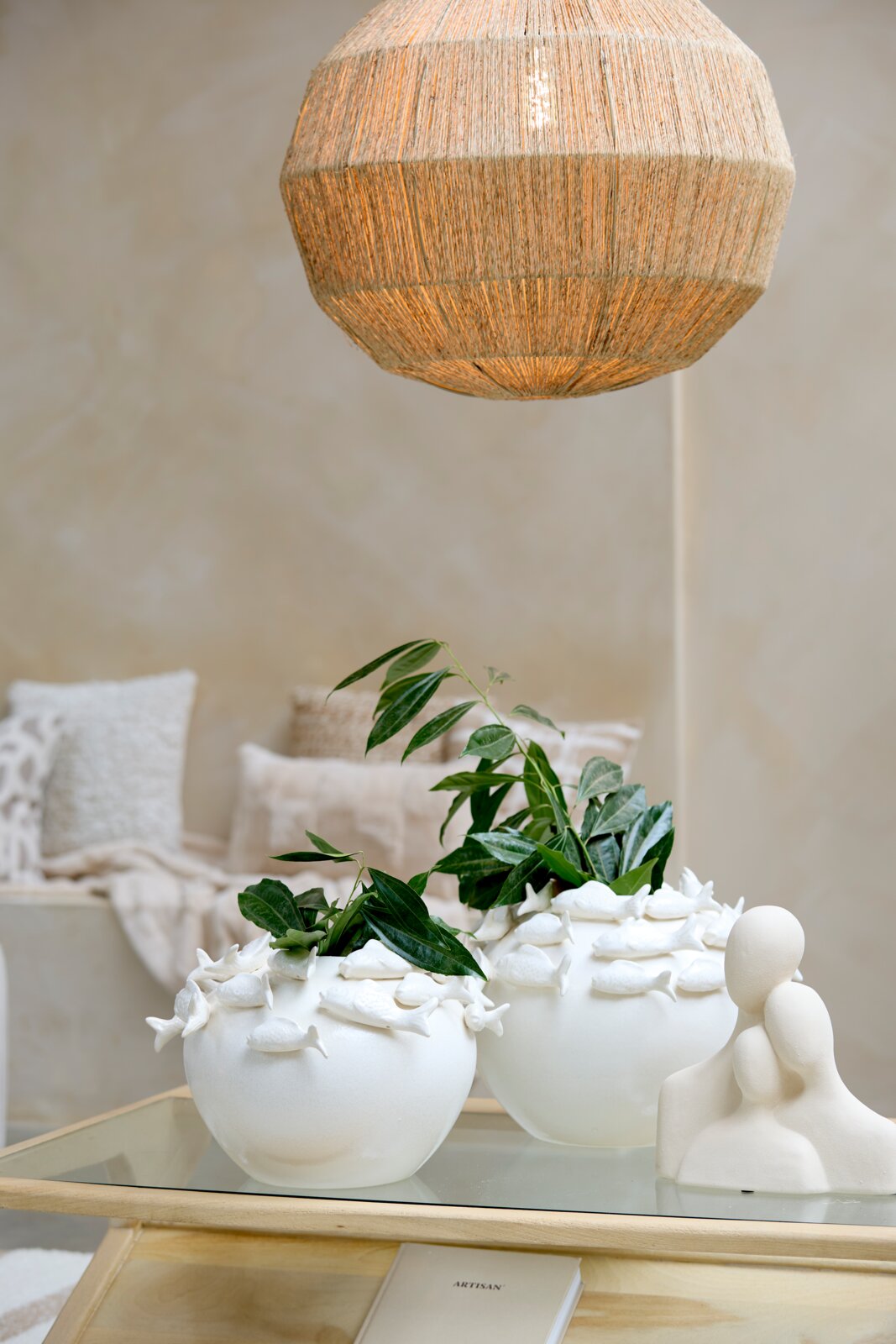 Light&Living Ornament 29cm SOLINE Zwei weiße Vasen mit Fischornamenten, gefüllt mit grünen Pflanzen, auf einem Glastisch. Im Hintergrund eine gemütliche Wohnzimmerszene mit Kissen und einer hängenden Lampe. Perspektive: seitlich.
