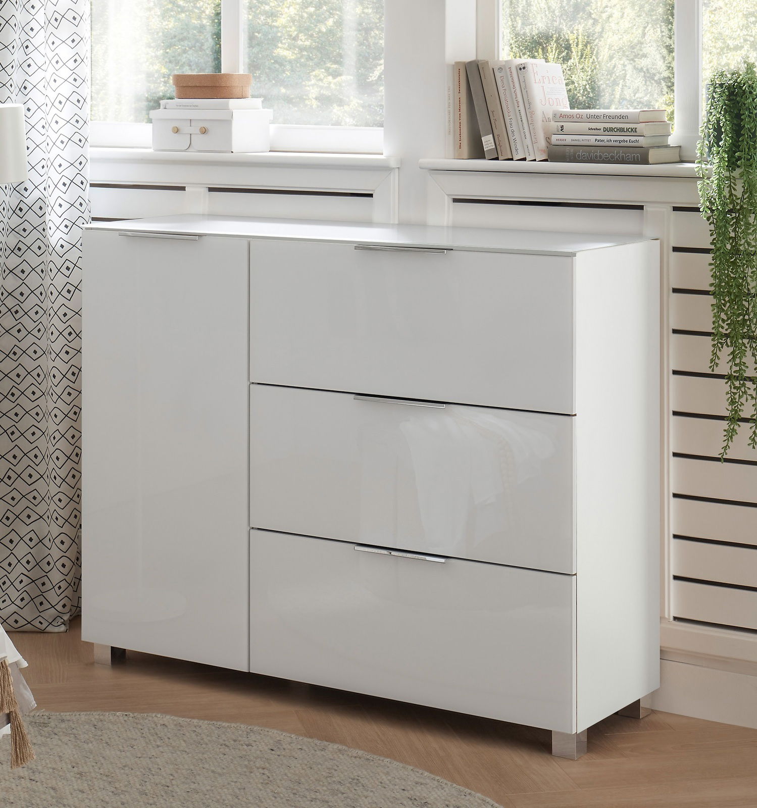 Weiße, moderne Kommode mit Hochglanzoberfläche, drei Schubladen und zwei Türen, seitliche Perspektive im Wohnzimmer.