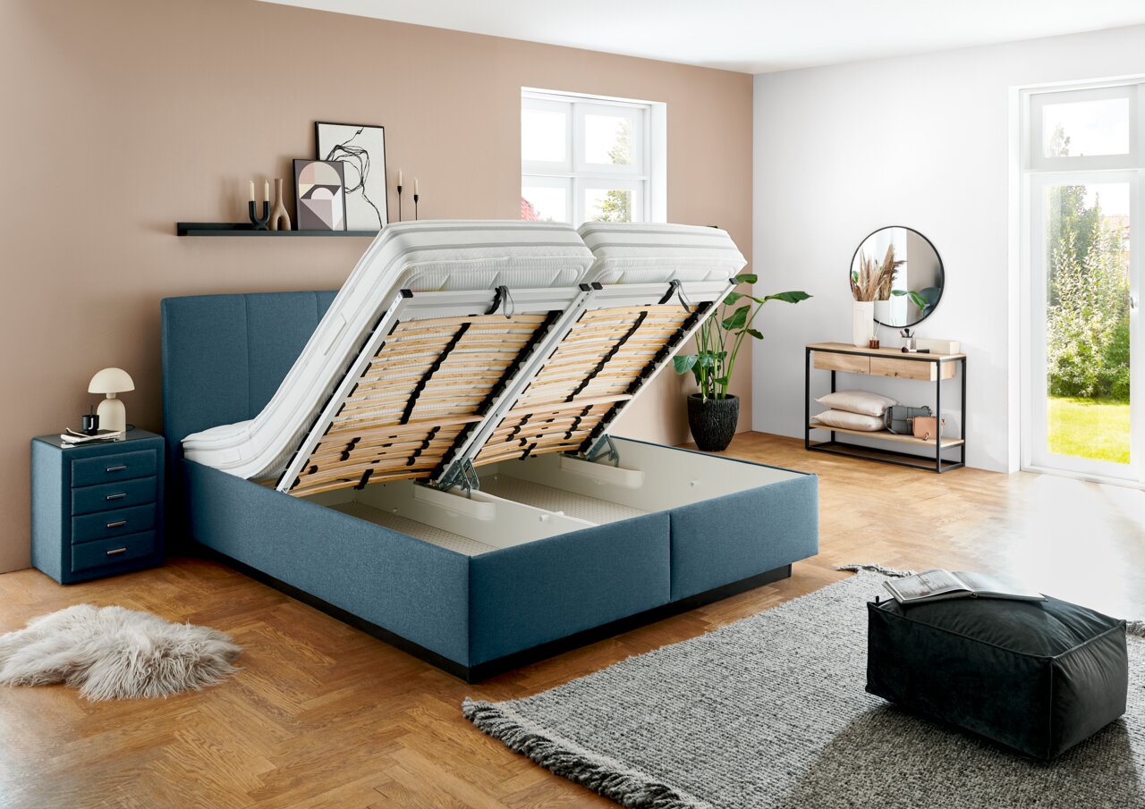 Modernes Polsterbett mit hochgeklappten Matratzen, das großzügigen Stauraum darunter zeigt. Perspektive: seitliche Ansicht in einem stilvoll eingerichteten Schlafzimmer.