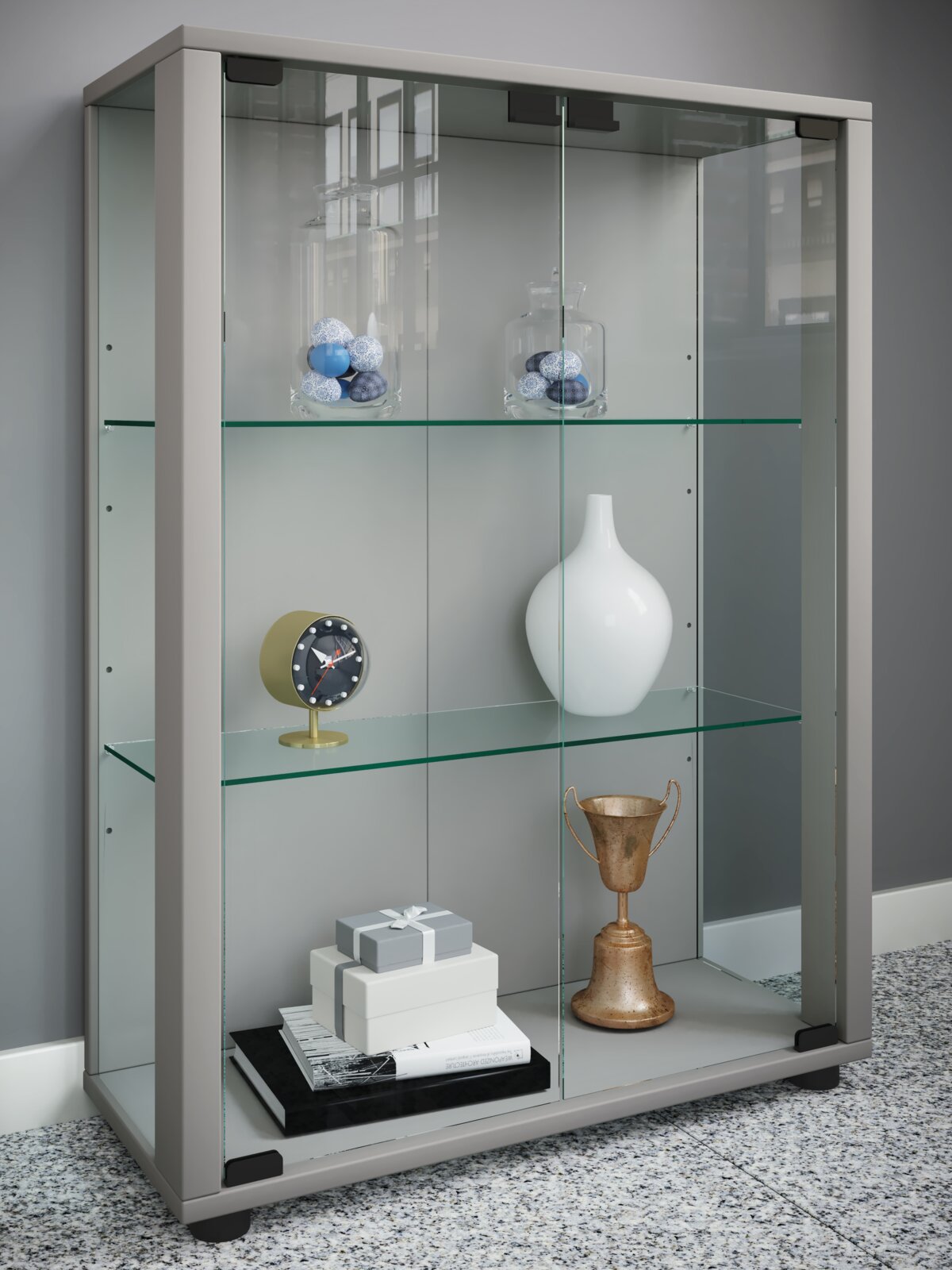 Moderne Glasvitrine mit grauem Rahmen und zwei Glasböden, bestückt mit Deko (Vasen, Uhr, Pokal, Boxen), in leichter Frontansicht von links