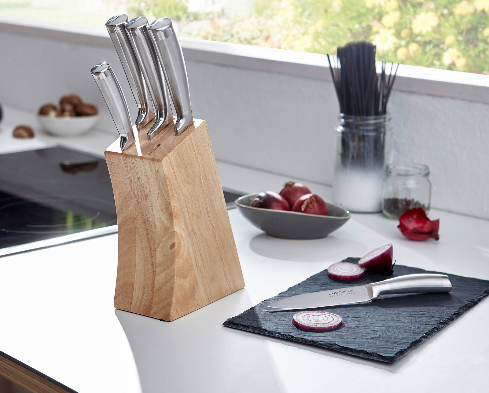 Messerblock aus Holz mit sechs Edelstahlmessern, auf einer Küchenarbeitsplatte platziert, mit einem geschnittenen Messer und Zwiebeln im Vordergrund. Perspektive von der Seite.