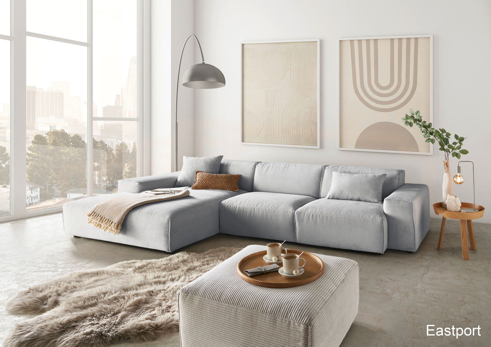 Lounge Handmade Ecksofa LH-EDINA Moderne Wohnzimmeransicht mit einer großen, hellgrauen Eckcouch, einem passenden Hocker und einem runden Holztisch. Im Hintergrund sind große Fenster mit Stadtblick und zwei abstrakte Kunstwerke an der Wand zu sehen.