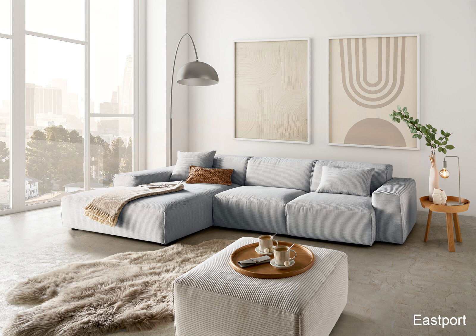 Lounge Handmade Ecksofa LH-EDINA Moderne Wohnzimmeransicht mit einer großen, hellgrauen Eckcouch, einem passenden Hocker und einem runden Holztisch. Im Hintergrund sind große Fenster mit Stadtblick und zwei abstrakte Kunstwerke an der Wand zu sehen.