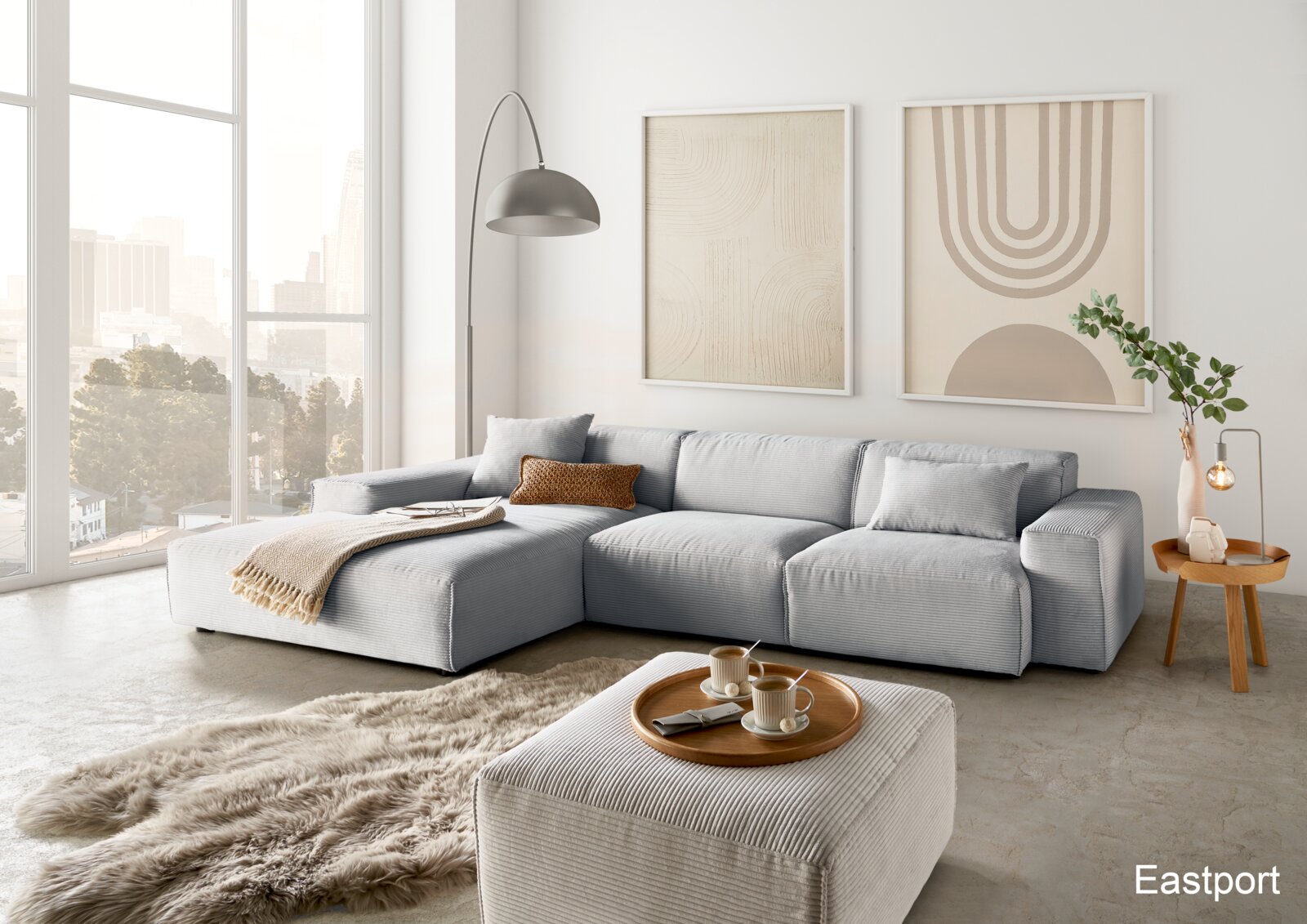 Lounge Handmade Hoekbank LH-EDINA Moderne Wohnzimmeransicht mit einer großen, hellgrauen Eckcouch, einem passenden Hocker und einem runden Holztisch. Im Hintergrund sind große Fenster mit Stadtblick und zwei abstrakte Kunstwerke an der Wand zu sehen.
