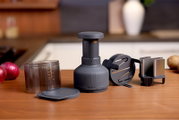 Schuin vooraanzicht van een grijze Zyliss handmatige foodprocessor/uienhakker met maatbeker, deksel en snij-inzetstukken op een houten aanrecht in een keukenomgeving.