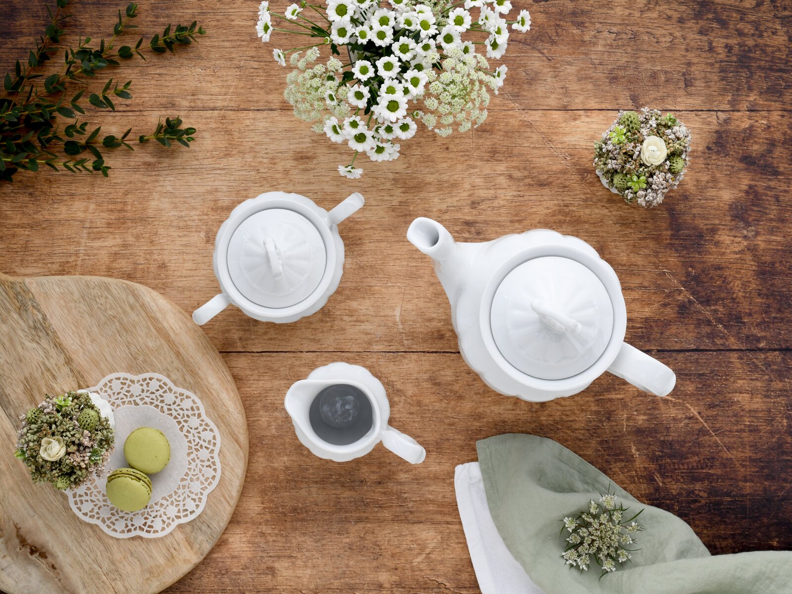 Draufsicht auf ein weißes Porzellan-Teeservice mit zwei Teekannen und einem Milchkännchen auf rustikalem Holztisch, dekoriert mit weißen Blumen und grünen Macarons auf einem Holzbrett