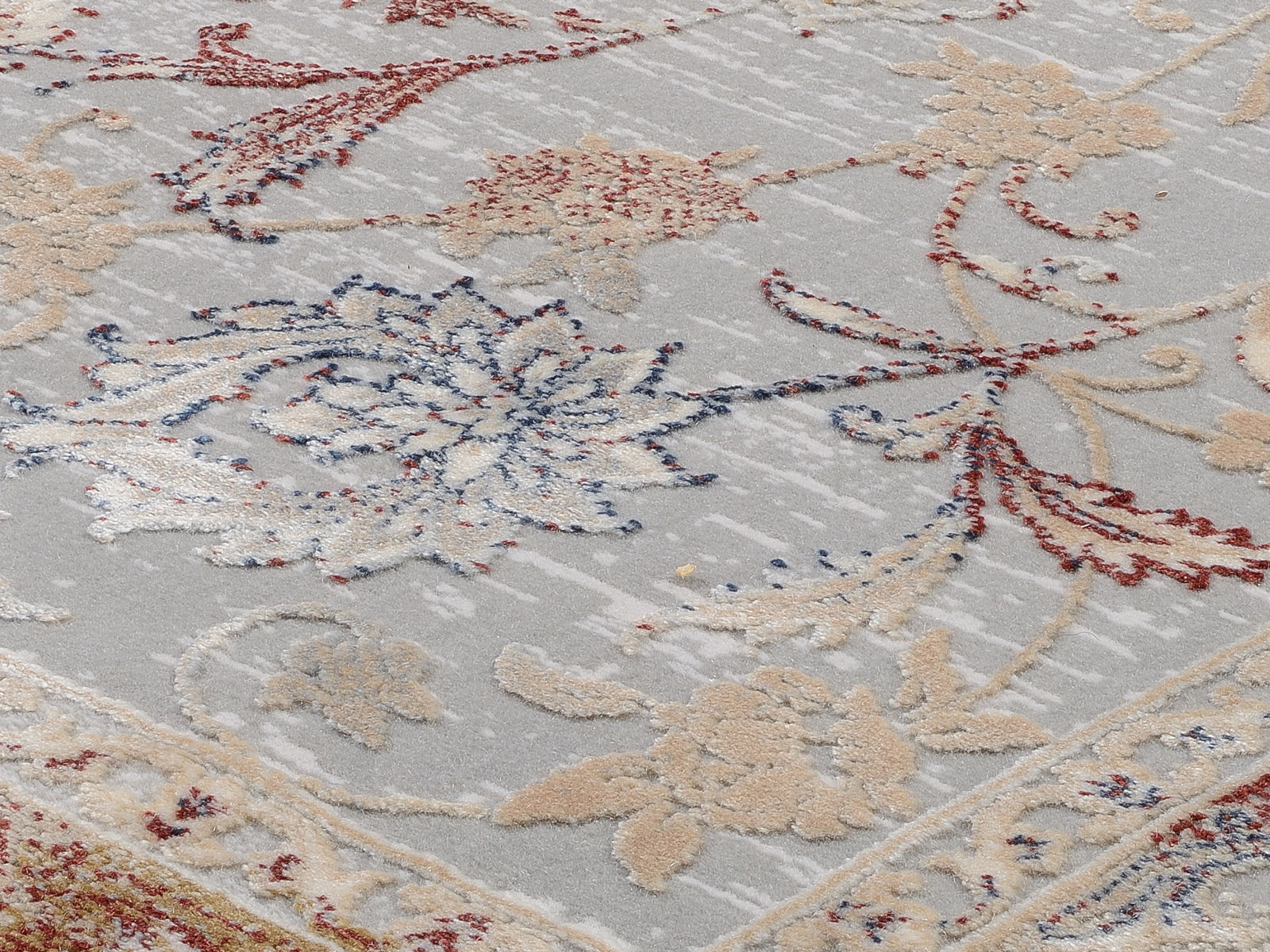 Nahaufnahme eines Orientteppichs mit floralen Mustern in Rot, Blau und Beige auf grauem Hintergrund, von schräg oben fotografiert.