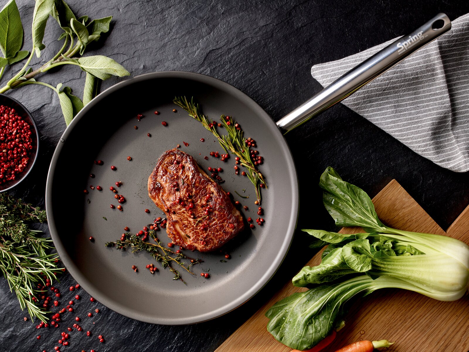 Bovenaanzicht van een grijze anti-aanbak koekenpan met roestvrijstalen handgreep (Spring), met een gebakken steak, rozemarijn en rode peperkorrels op een donkere stenen ondergrond, naast verse kruiden en groenten.