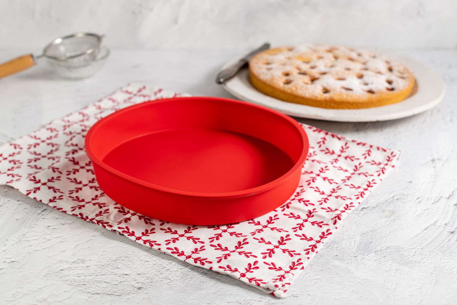Rote Silikon-Rundform mit einem Durchmesser von 26 cm, auf einem dekorativen Tuch in der Küche platziert, seitliche Perspektive.