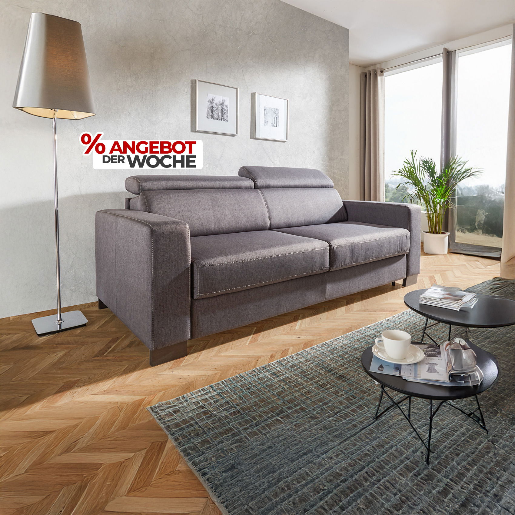 Graues 3-Sitzer Sofa mit Kopfstützen im Wohnzimmer neben Stehlampe, Couchtisch und Fensterfront