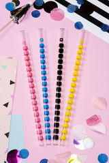 Vier bunte Glastrinkhalme in den Farben Pink, Blau, Schwarz und Gelb auf einem rosa Hintergrund, von oben betrachtet, umgeben von dekorativen Papierkonfetti.