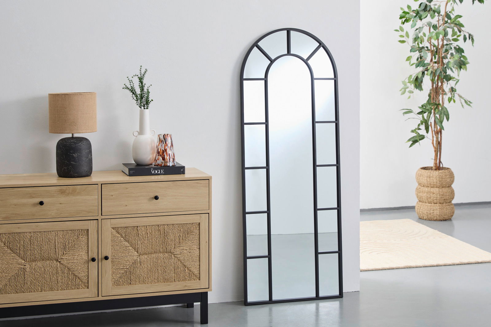 Landscape Spiegel ca. 61x161 cm LAUREL Großer, rechteckiger Spiegel mit schwarzem Rahmen und Bogen an der Oberseite, an eine weiße Wand gelehnt, neben einer Holzkommode und einer Pflanze. Perspektive von vorne.