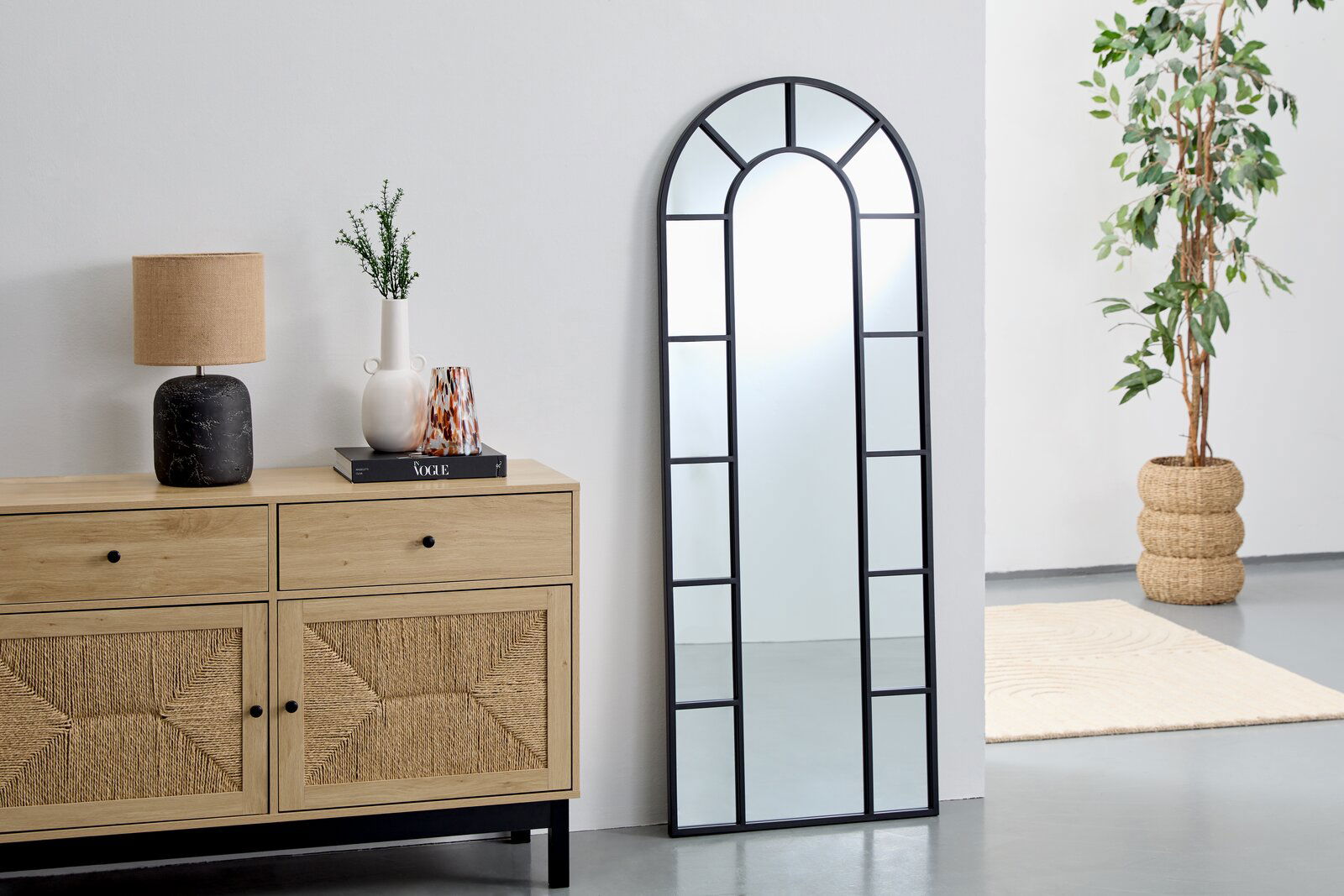 Landscape Spiegel ca. 61x161 cm LAUREL Großer, rechteckiger Spiegel mit schwarzem Rahmen und Bogen an der Oberseite, an eine weiße Wand gelehnt, neben einer Holzkommode und einer Pflanze. Perspektive von vorne.