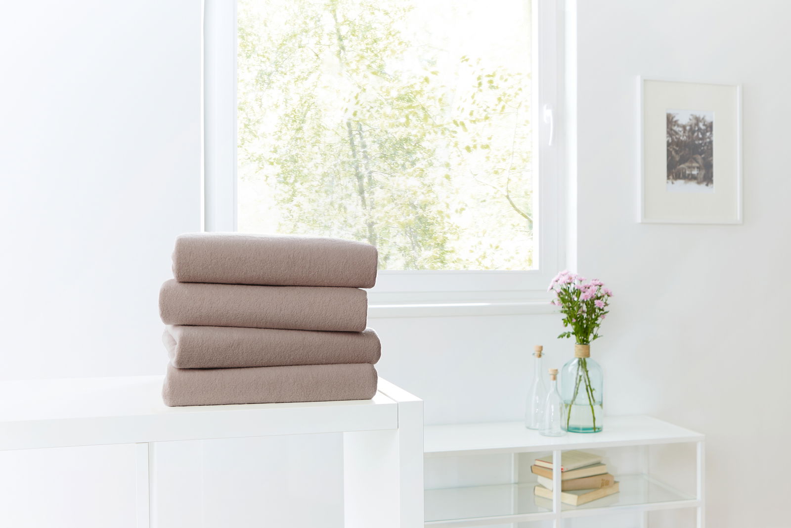Drei gefaltete Spannbetttücher in Beige auf einem weißen Tisch, im Hintergrund ein Fenster mit Blick ins Grüne.