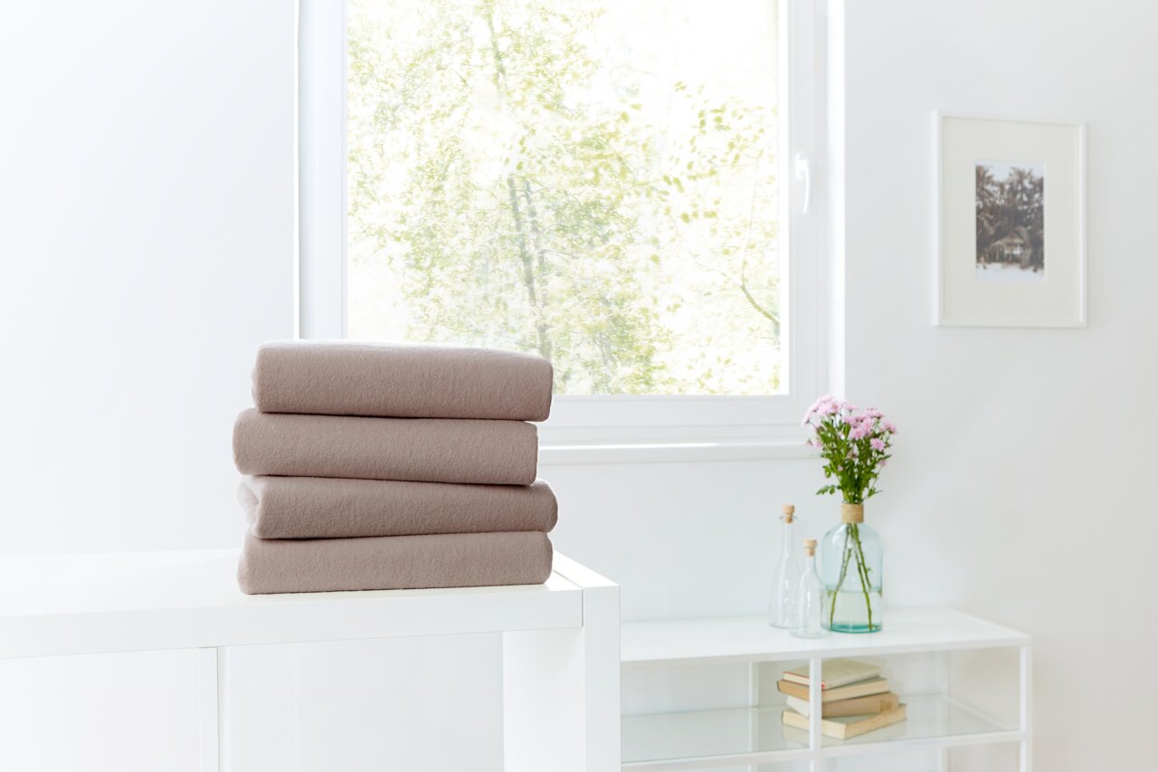 Novel Spannbetttuch TEDDY-FLAUSCH Drei gefaltete Spannbetttücher in Beige auf einem weißen Tisch, im Hintergrund ein Fenster mit Blick ins Grüne.