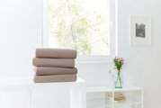 Drei gefaltete Spannbetttücher in Beige auf einem weißen Tisch, im Hintergrund ein Fenster mit Blick ins Grüne.