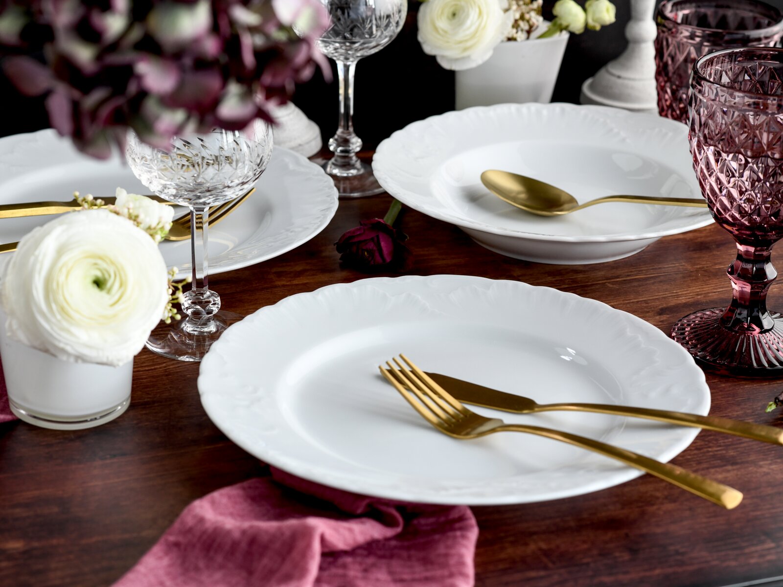 Festlich gedeckter Holztisch mit weißen Porzellantellern, goldenem Besteck, Kristall- und rosa Gläsern sowie Blumen, aus leicht schräger Seitenansicht