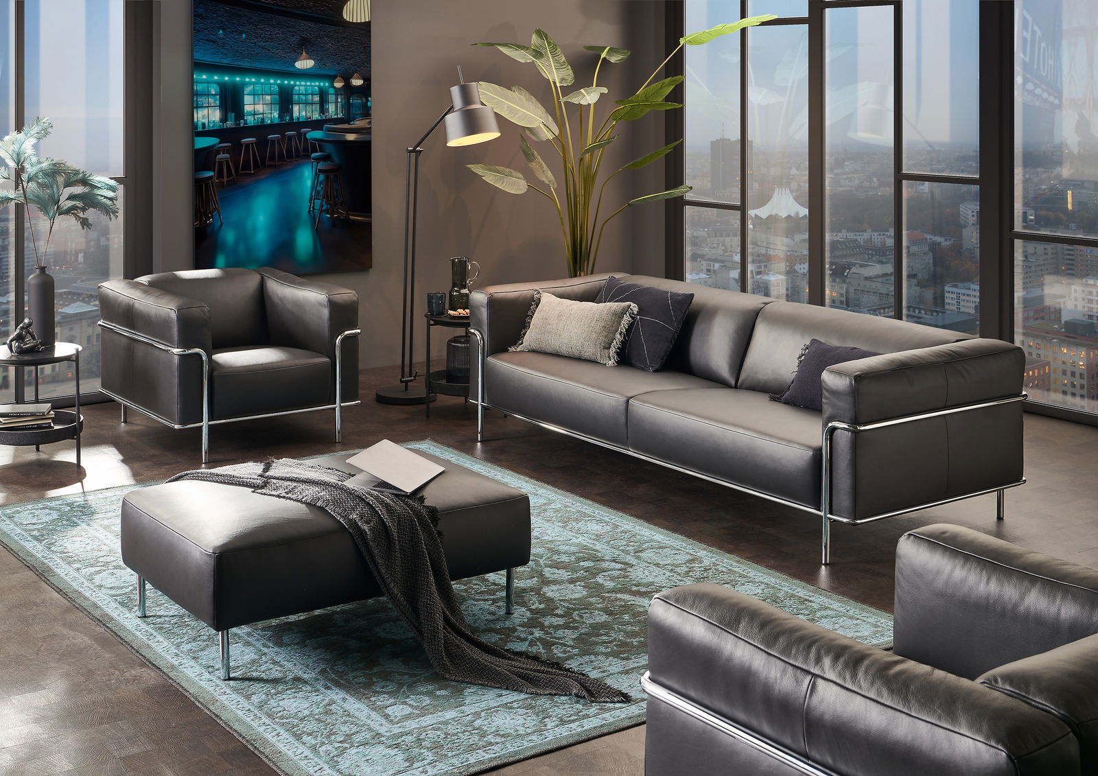 Moderne Wohnzimmermöbel in einem stilvollen Raum mit einem großen Fenster im Hintergrund, bestehend aus einem dreisitzigen Sofa, einem Sessel und einem Hocker in schwarzem Leder mit Metallrahmen, aus der Perspektive von vorne rechts betrachtet.