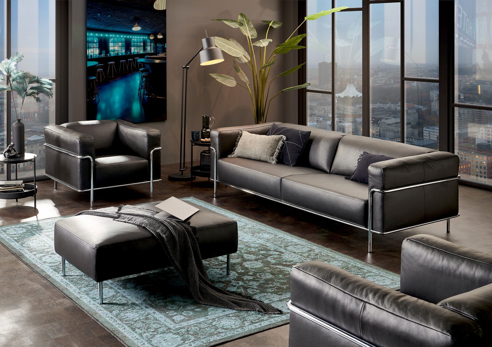 Moderne Wohnzimmermöbel in einem stilvollen Raum mit einem großen Fenster im Hintergrund, bestehend aus einem dreisitzigen Sofa, einem Sessel und einem Hocker in schwarzem Leder mit Metallrahmen, aus der Perspektive von vorne rechts betrachtet.
