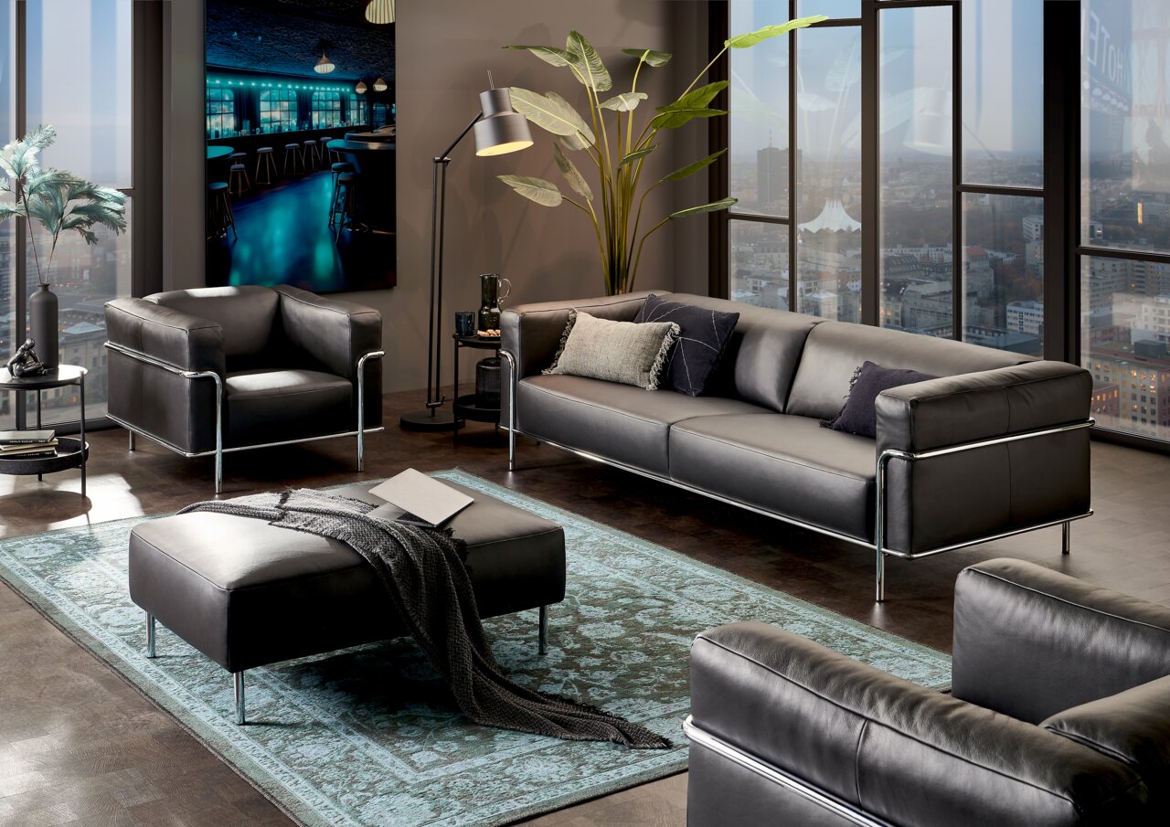 Johann Jakob Fauteuil MANDIA Moderne woonkamermeubels in een stijlvolle ruimte met een groot raam op de achtergrond, bestaande uit een driezitsbank, een fauteuil en een poef van zwart leer met een metalen frame, gezien vanuit het perspectief rechtsvoor.