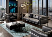 Moderne Wohnzimmermöbel in einem stilvollen Raum mit einem großen Fenster im Hintergrund, bestehend aus einem dreisitzigen Sofa, einem Sessel und einem Hocker in schwarzem Leder mit Metallrahmen, aus der Perspektive von vorne rechts betrachtet.