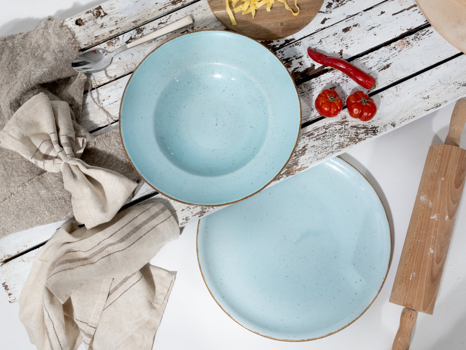 Zwei hellblaue, gesprenkelte Keramik-Teller/Schalen mit braunem Rand auf rustikalem, weißem Holztisch mit Küchenaccessoires, Draufsicht (Top-Down).
