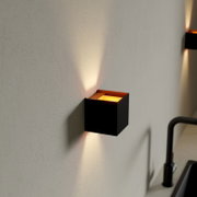 Schwarze, würfelförmige Wandleuchte mit warmweißem Up-&-Down-Lichtkegel an heller Wand, Perspektive: seitliche Nahaufnahme leicht von vorn