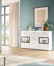 Moderne weiße Hochglanz-Kommode/Sideboard mit sechs Schubladen und zwei Glas-Displayfächern, dekoriert mit Büchern, Vase und Pflanze, in einem hellen Wohnzimmer; Perspektive: frontale Ansicht leicht von links.