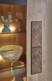 Detailansicht eines modernen Holz-Vitrinenschranks mit Glasböden und warmem Innenlicht; sichtbar sind Weingläser, eine dekorative Schale und eine Front mit zwei rechteckigen Holz-Einlegeelementen in Baumring-Optik, fotografiert in schräger Nahaufnahme von