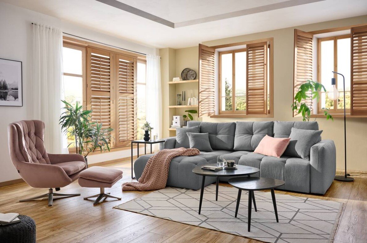 Gezellige zithoek met een grijze hoekbank, een roze fauteuil en een poef, aangevuld met moderne houten meubels en planten. Aanzicht van voren.