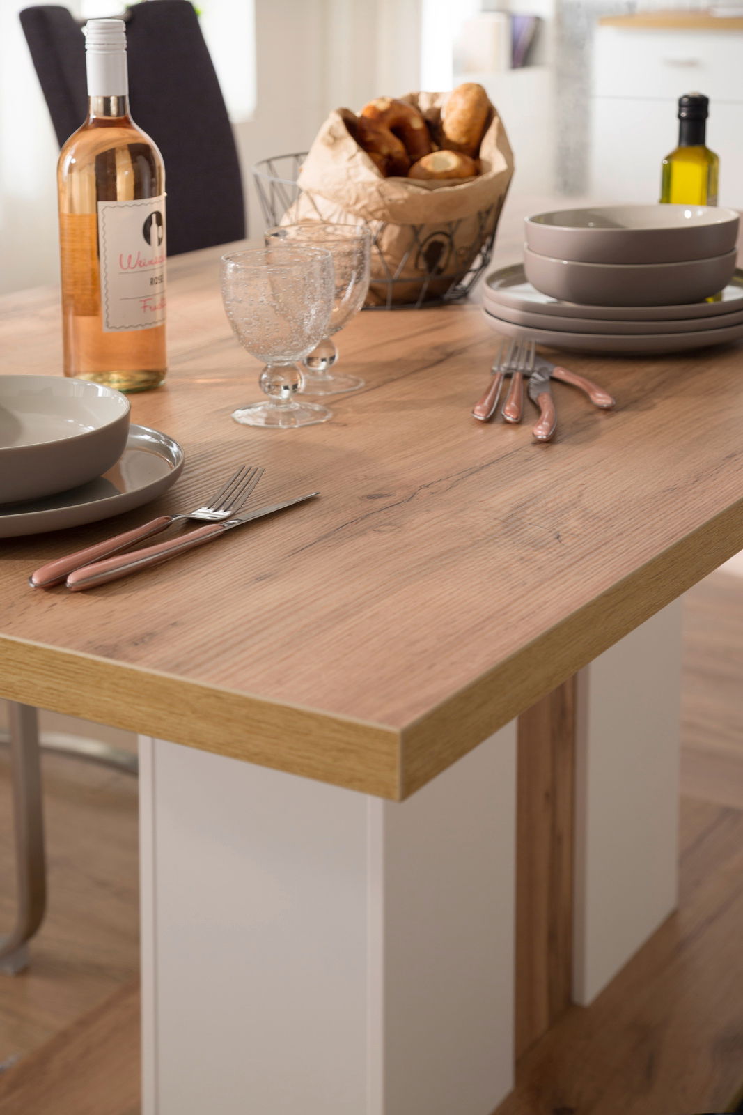 Nahaufnahme eines Esstisches mit Holzoberfläche, gedeckt mit Geschirr, Besteck, einer Flasche Wein und einem Brotkorb.