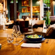 Salatschüssel auf blauem Teller mit Besteck, Wasserglas und Weißweinglas auf Holztisch in gemütlichem Restaurant, aufgenommen aus Tischhöhe in leicht schräger Vorderansicht