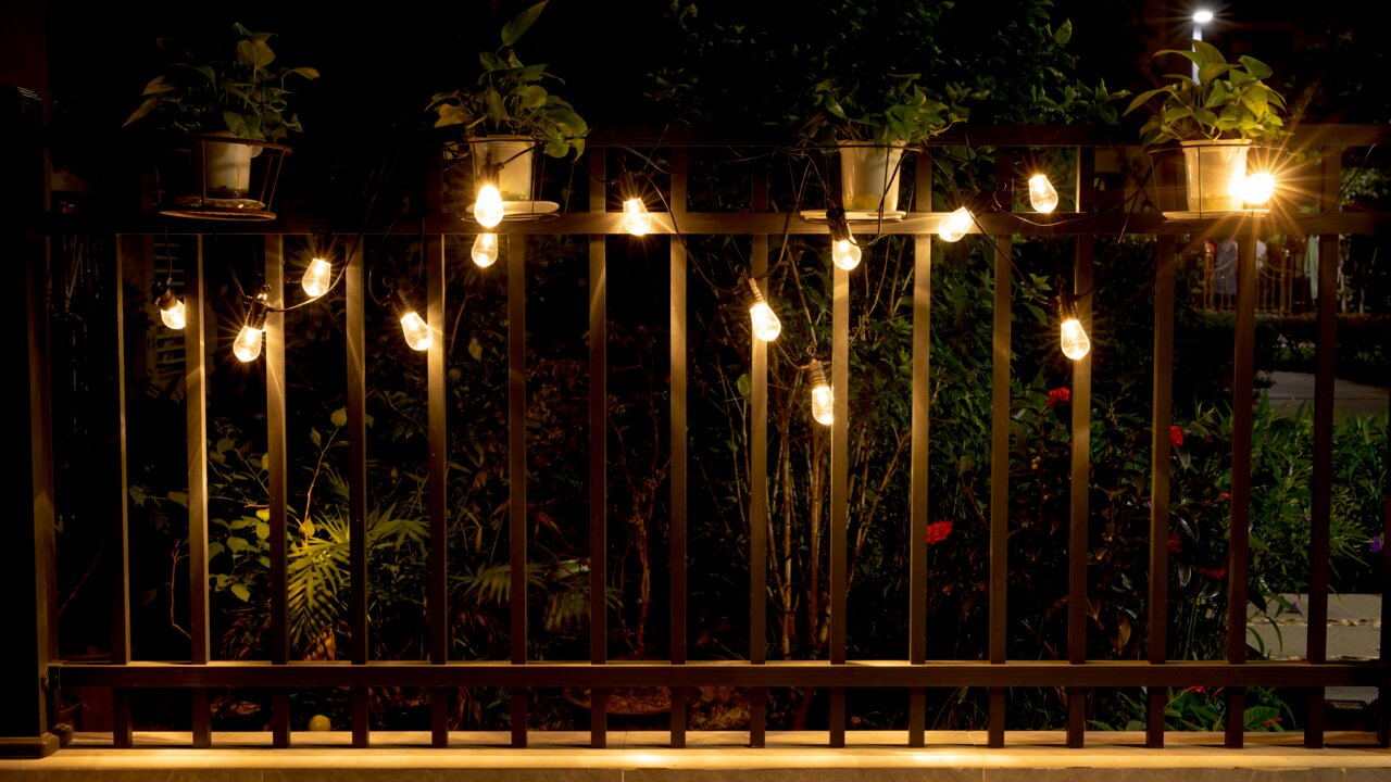 Een lichtslinger op een balkon, opgehangen aan een reling met planten op de achtergrond, 's nachts gefotografeerd.