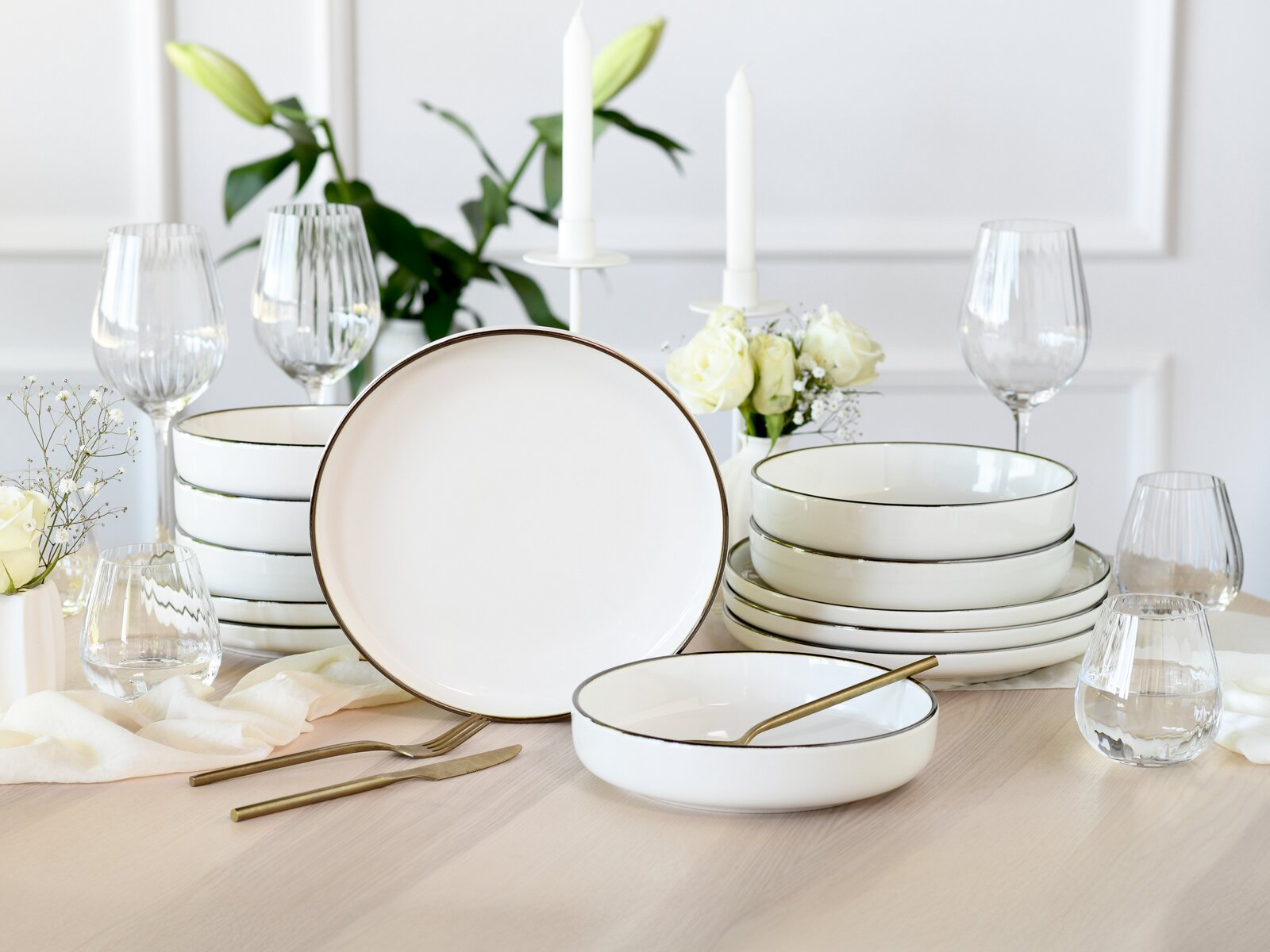 Elegantes weißes Porzellan-Geschirrset mit feinem Goldrand, Teller und Schalen auf gedecktem Tisch mit Gläsern, Kerzen und weißen Blumen, frontal in leichter Schrägansicht aufgenommen