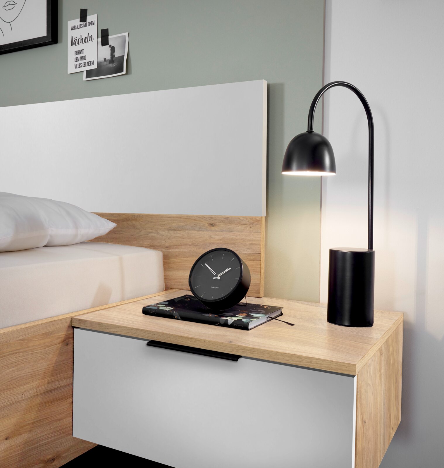 Nachtkastje in hout en wit, met daarop een zwarte tafellamp en een zwarte wekker, gezien vanaf de zijkant