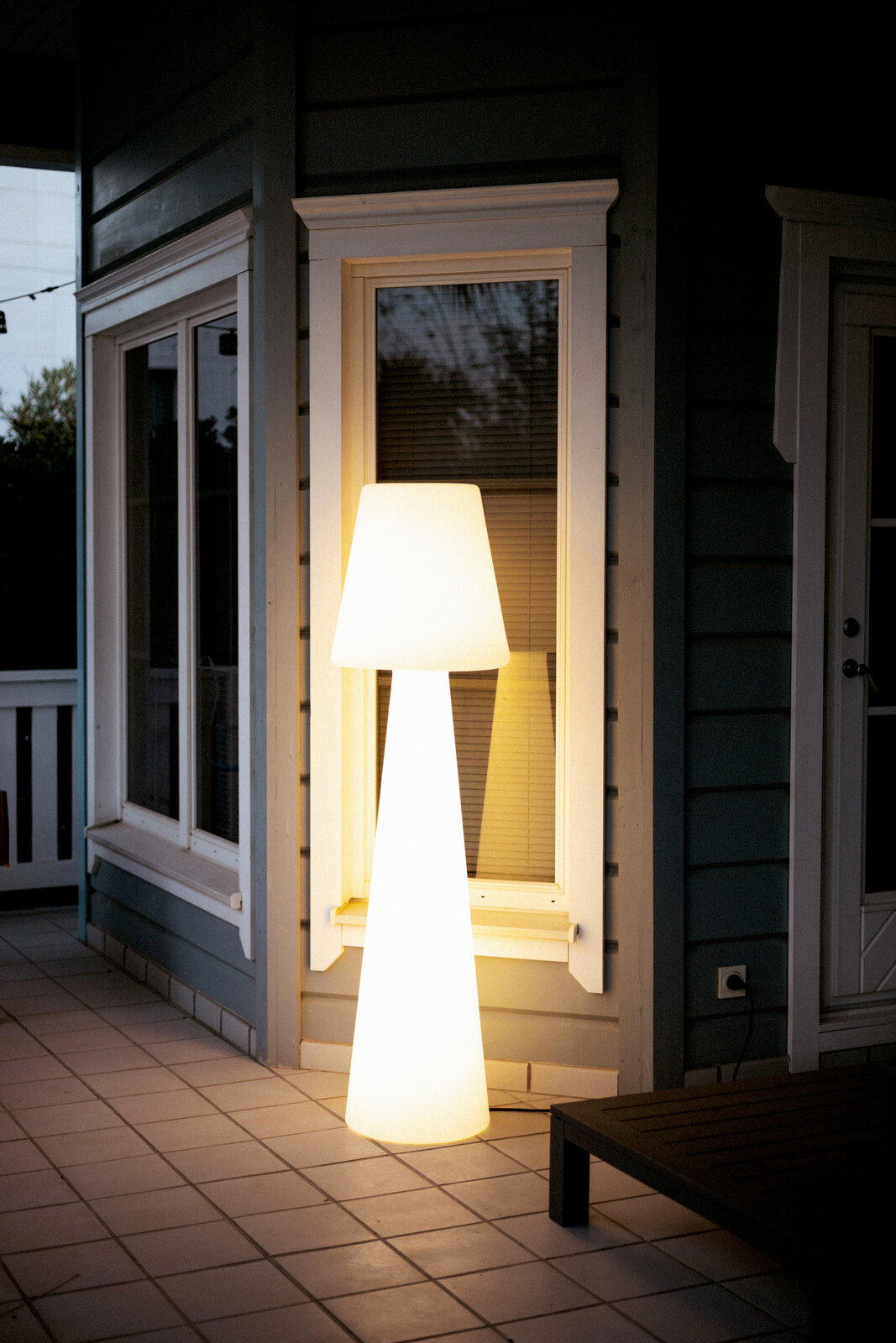 Leuchtende, moderne Outdoor-Stehleuchte in Weiß mit konischem Schirm und schlankem Korpus auf einer gefliesten Terrasse vor einem Hausfenster, aufgenommen in leichter Seitenansicht bei Dämmerung.