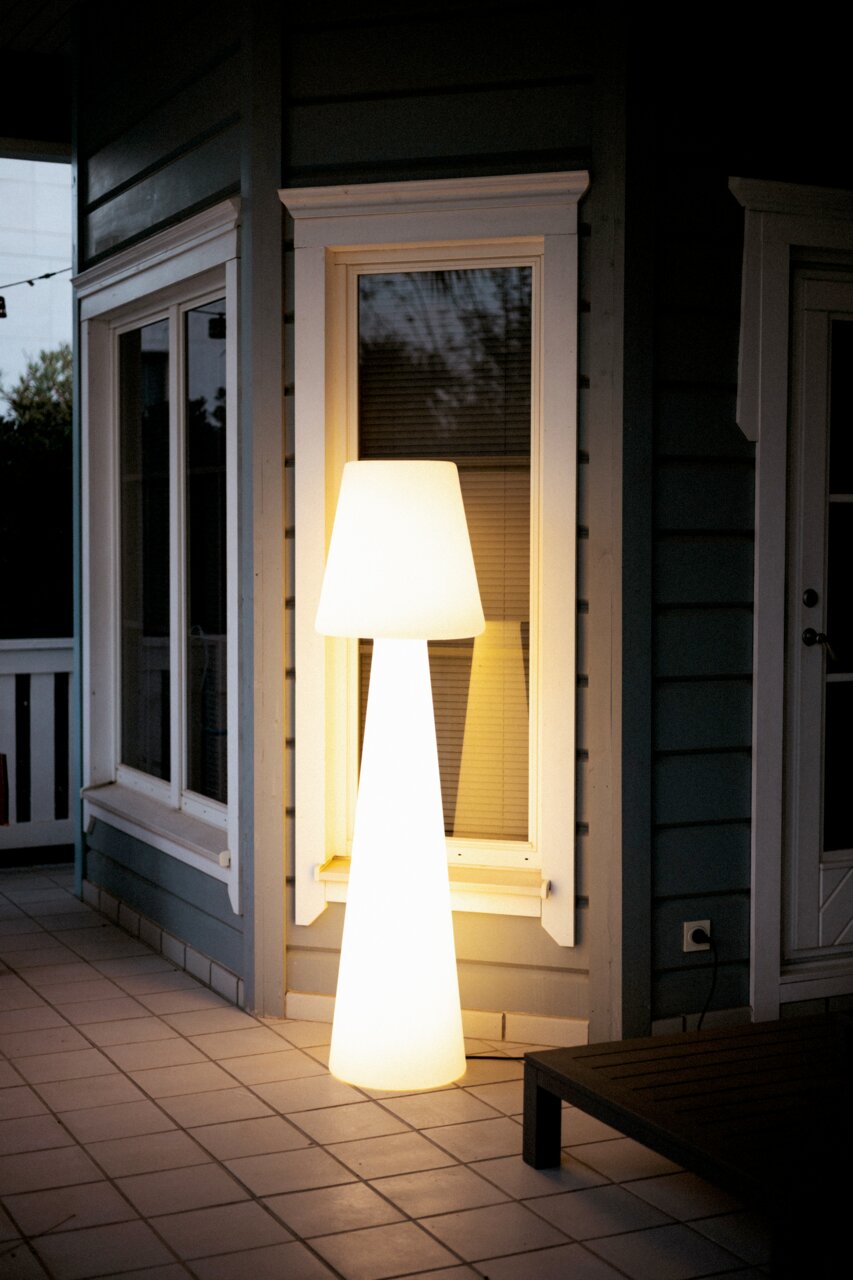 Een stralende, moderne buitenstaande lamp in wit met een conische kap en een slanke voet, op een betegeld terras voor een raam van een huis, gefotografeerd in een licht zijaanzicht bij schemering.