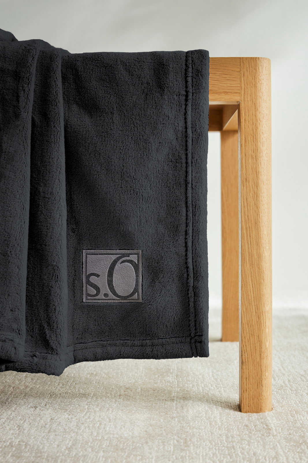 Schwarze s.Oliver Kuscheldecke aus weichem Material, drapiert über eine Holzkante, mit sichtbarem Logo. Perspektive von der Seite.