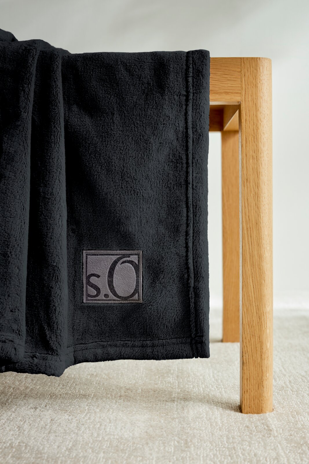 Schwarze s.Oliver Kuscheldecke aus weichem Material, drapiert über eine Holzkante, mit sichtbarem Logo. Perspektive von der Seite.