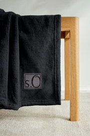 Schwarze s.Oliver Kuscheldecke aus weichem Material, drapiert über eine Holzkante, mit sichtbarem Logo. Perspektive von der Seite.