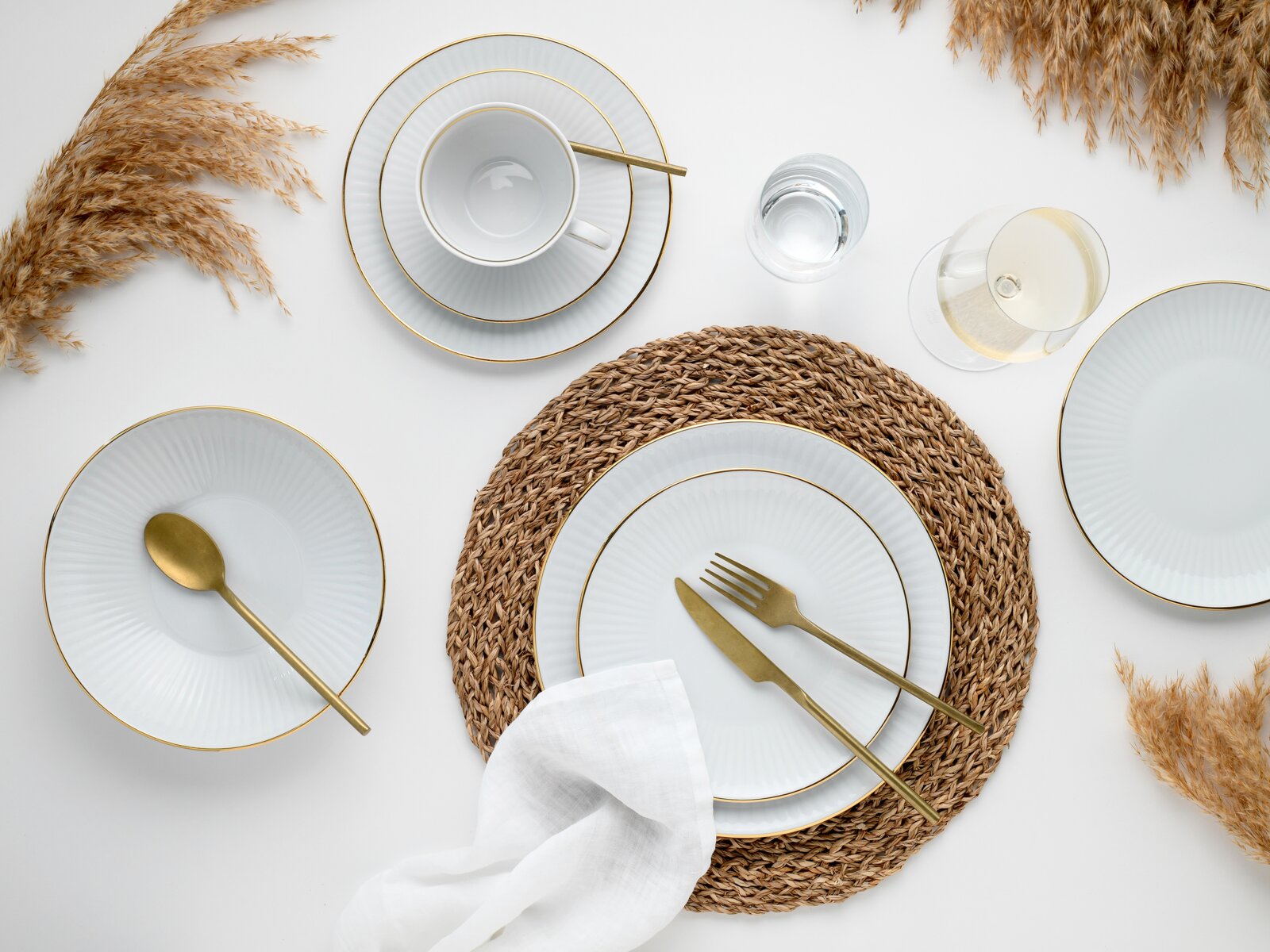 Draufsicht auf ein elegant gedecktes Tischarrangement mit weißem Porzellan-Geschirr mit Goldrand, goldfarbenem Besteck, geflochtenem Platzset, Wasserglas und Weißweinglas auf hellem Hintergrund, dekoriert mit Trockengras.