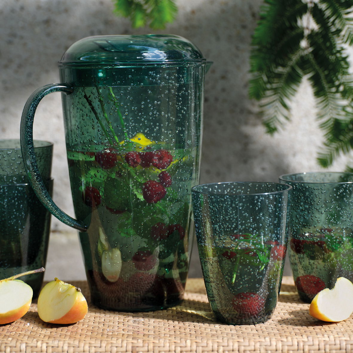Grünes Kannenset mit Bechern, gefüllt mit Wasser, Himbeeren und Minzblättern, auf einem Tisch mit Apfelstücken, seitliche Perspektive.