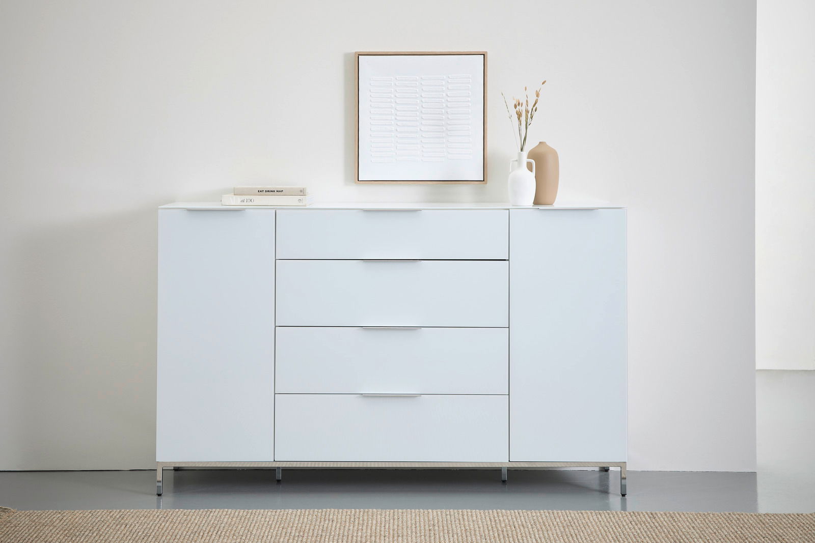 Modernes weißes Sideboard mit vier Schubladen und zwei Türen, dekoriert mit Büchern und Vasen, aus frontaler Perspektive.