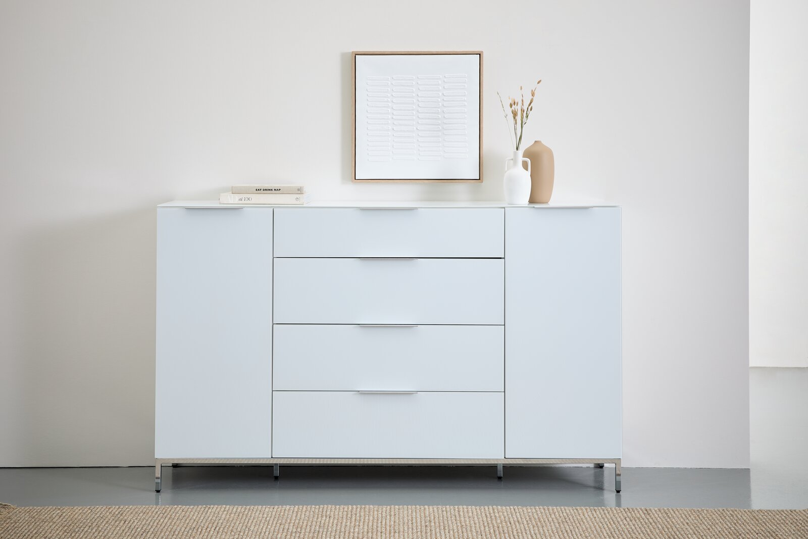 Modernes weißes Sideboard mit vier Schubladen und zwei Türen, dekoriert mit Büchern und Vasen, aus frontaler Perspektive.