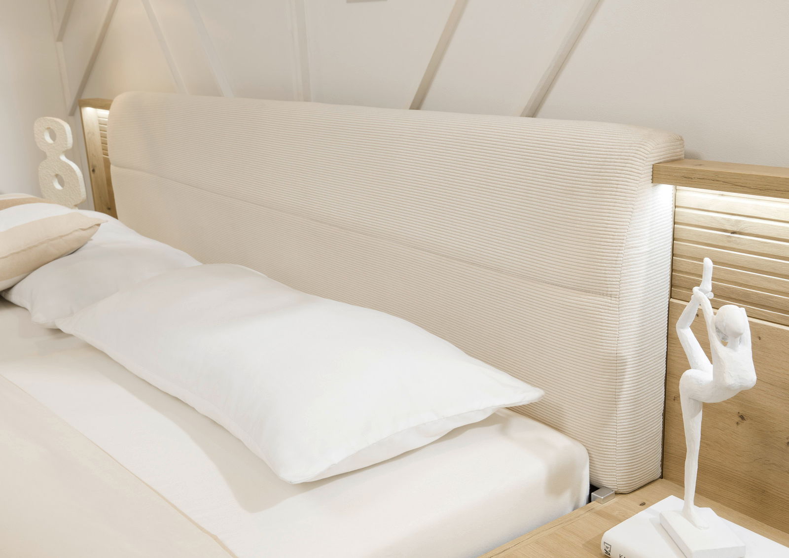 Nahaufnahme eines Bettes mit gepolstertem Kopfteil in Cremefarbe, flankiert von Holzelementen und dekorativer Beleuchtung. Perspektive von der Seite.