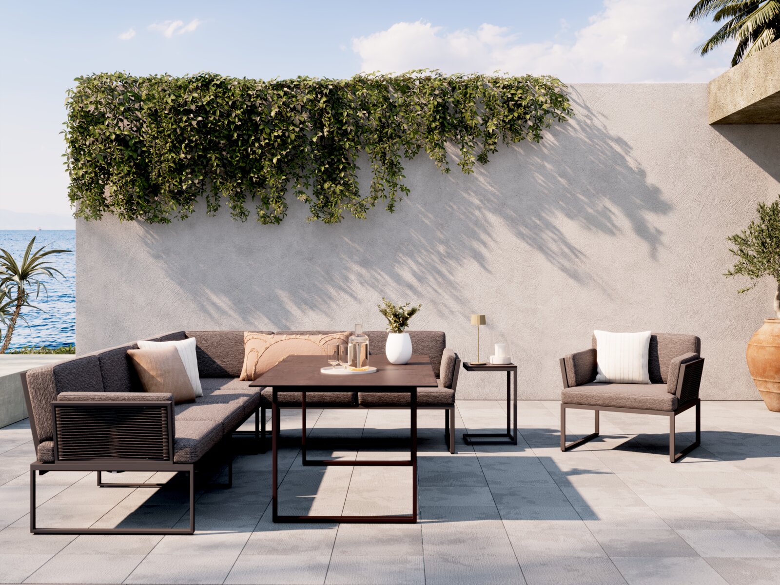 Amatio Loungetisch GIRONA Frontale Perspektive: moderne Outdoor-Lounge mit grauen Polstern, L-förmigem Sofa, passendem Sessel und niedrigem Couchtisch auf sonniger Terrasse vor heller Wand mit hängendem Grün und Meerblick.