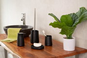 Schwarzer Seifenspender Kira auf einem Holzwaschtisch, umgeben von weiteren schwarzen Badaccessoires und einer Pflanze, aus seitlicher Perspektive fotografiert.